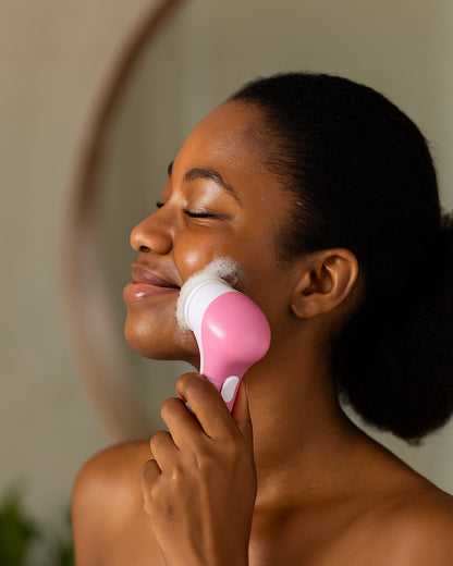 Woman using a pink 5-in-1 facial cleansing brush on her face in the bathroom